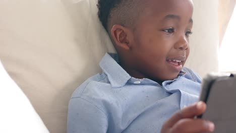 happy african american boy lying on sofa using tablet at home, slow motion