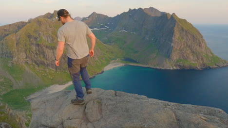 Tourist-walking-towards-the-worlds-famous-edge-on-the-Peak-of-Ryten,-with-kvalvika-beach,-Lofoten-Island,-Norway