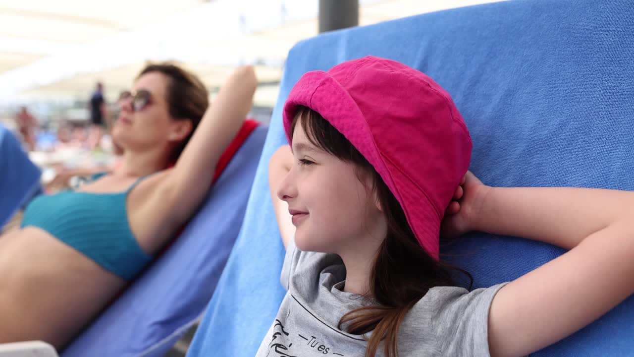 Girl in pink panama rests lying on sun lounger near mother