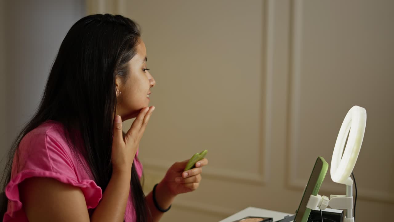 On the side a happy brunette Indian girl does a facial massage using a special massager and talks about it during her Beauty blog about beauty and make-up in a modern apartment during the day in front of a phone screen