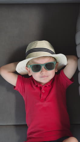 tired little boy in sunglasses and men straw hat lies on sofa near suitcase. funny toddler puts hands on head back looking in camera upper closeup