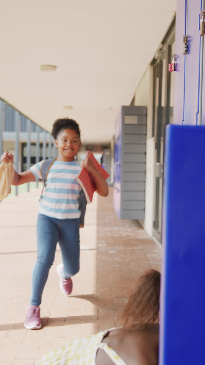 Vertical video of two happy, diverse schoolgirls talking by lockers in school corridor, copy space