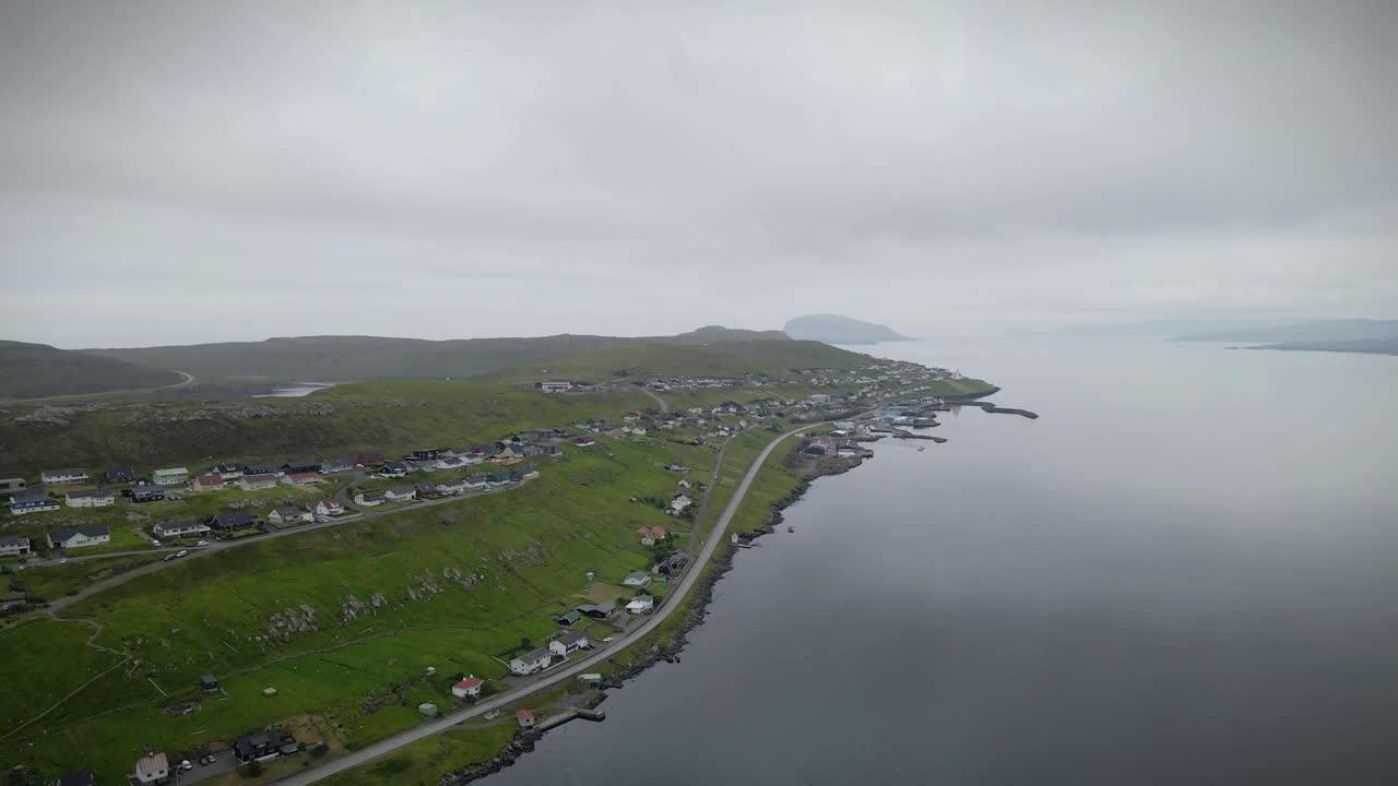 Drone capturing Toftir village in Eysturoy, Faroe Islands, showcasing houses along the coastline, roads winding through green hills, and a cloudy sky creating a serene atmosphere, drone revealing shot