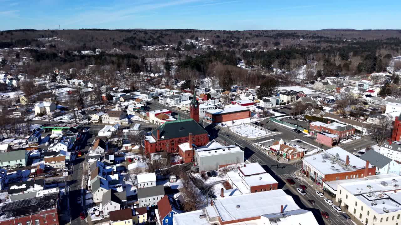 Vista aérea de Ware, Massachusetts, após uma nevasca, com igreja de tijolos vermelhos e ruas de cidade pequena cobertas por neve de inverno sob luz do dia brilhante. Tomada invertida.