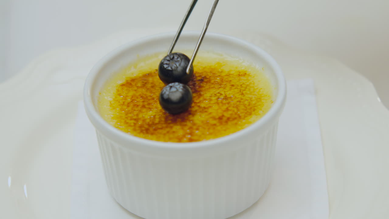 Close-up of a chef decorating a crispy creme brulee with blueberries using kitchen tongs