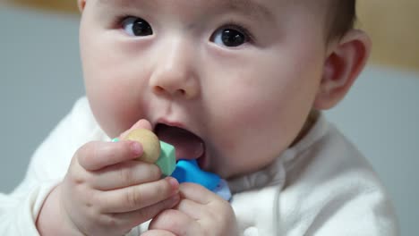 Lindo-Bebé-Pequeño-Masajeando-Su-Chicle-Tratando-De-Comer-Un-Brazalete-Colorido-Y-Mirando-A-La-Cámara,-Cara-De-Cerca