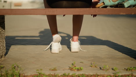 back leg view of lady removing left sneaker while sitting on bench under bright sunlight, casting soft shadows on pavement, with roller skate partially visible on bench