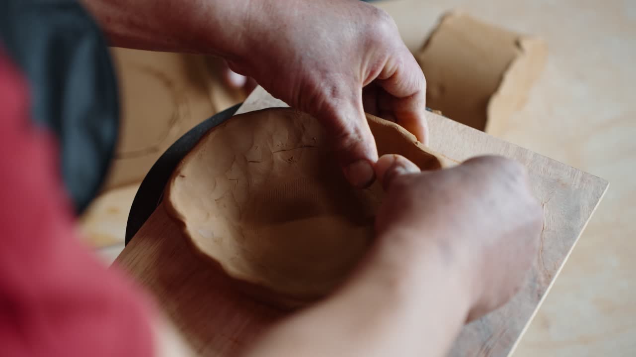 An over-the-shoulder close-up shot of an artisan's hands using a traditional hand-building technique to pinch and form a rustic clay bowl in a pottery workshop