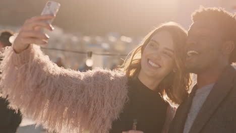 group of friends hanging out together enjoying rooftop party at sunset taking selfie photos using smartphone drinking alcohol having fun on weekend sharing celebration