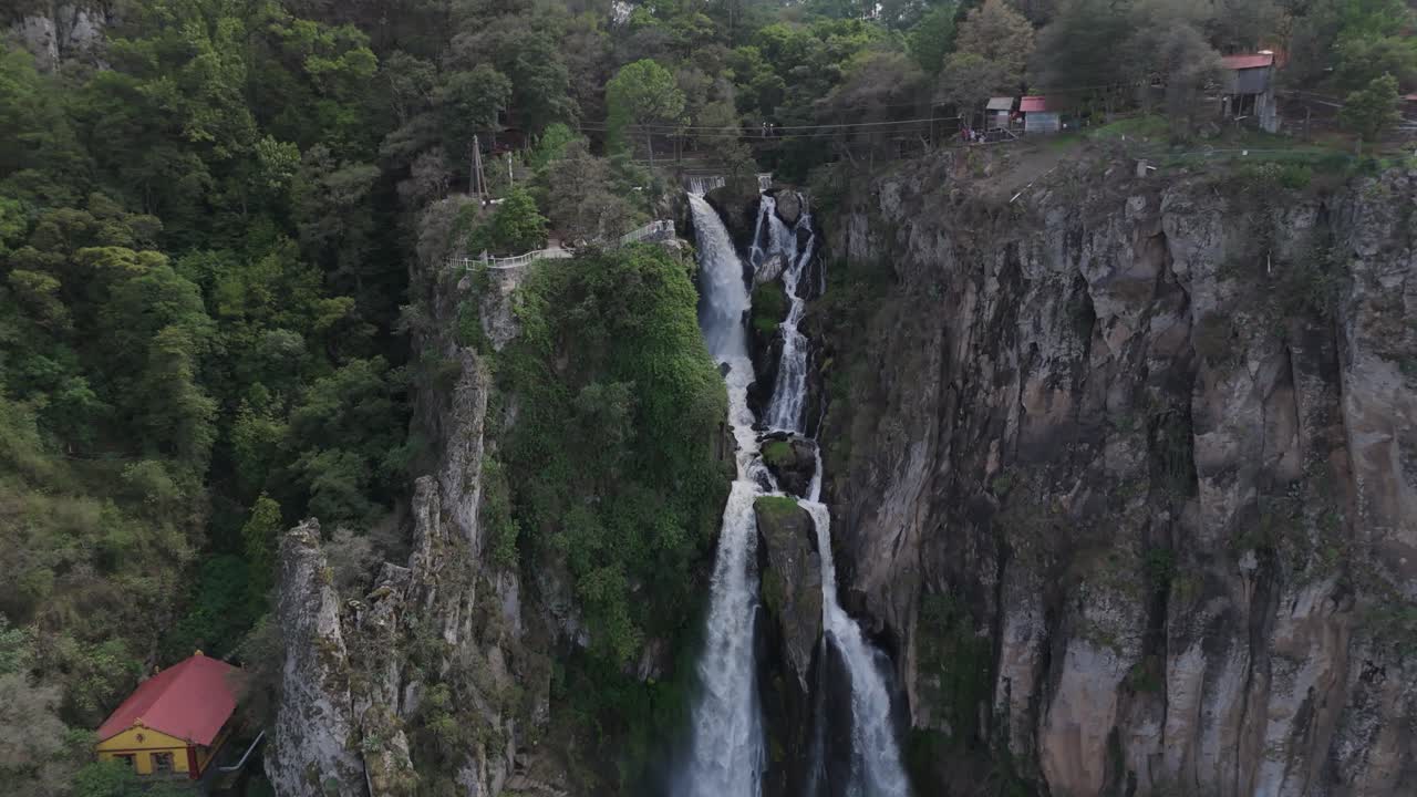 Aerial zoom-in reveals a massive waterfall surrounded by green rocky nature, with a few hidden houses tucked into the landscape.