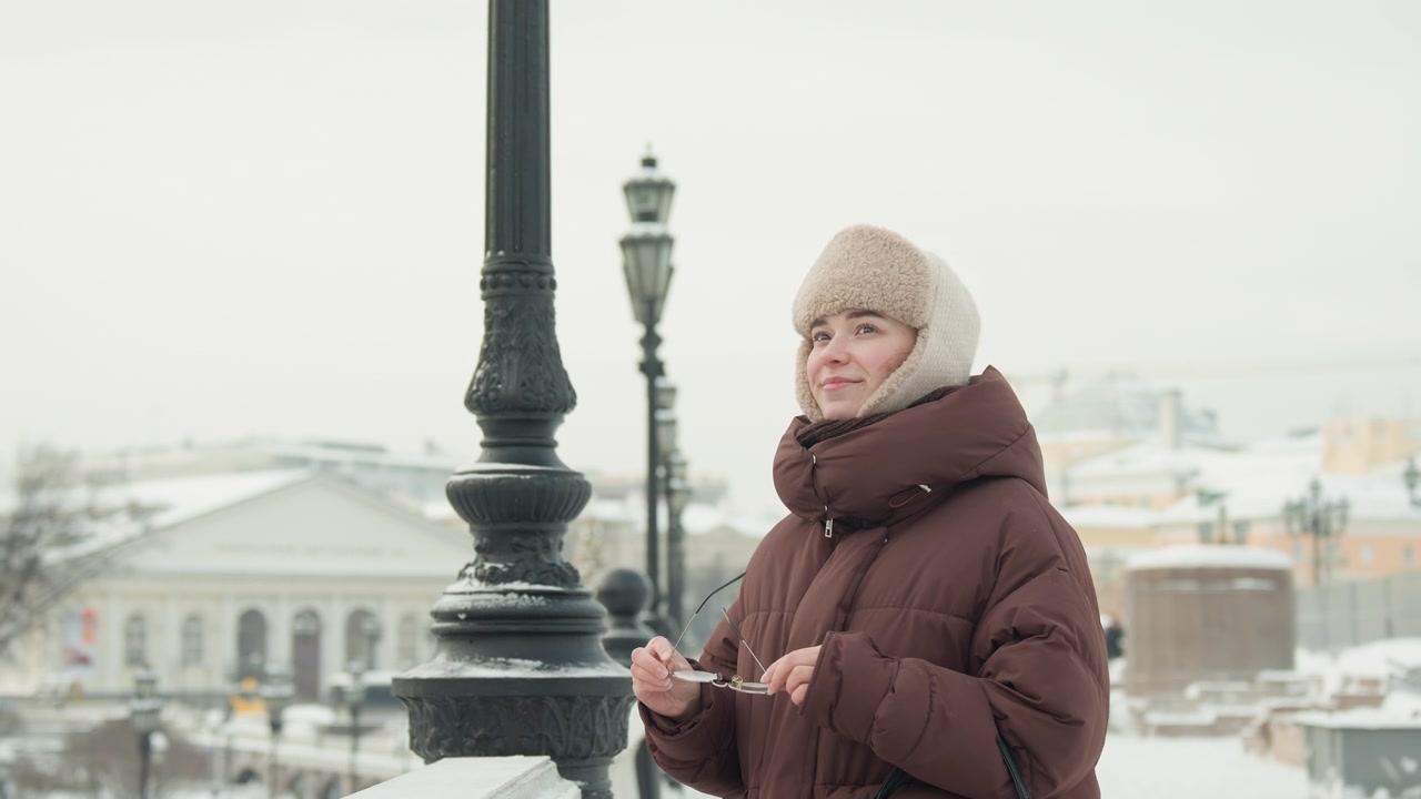 Donna infagottata con cappotto marrone e cappello di lana che tiene lo smartphone all'orecchio, aggiusta leggermente il cappello con la mano libera, in piedi in un parco cittadino innevato con edifici storici, lampioni e una soffusa luce invernale.