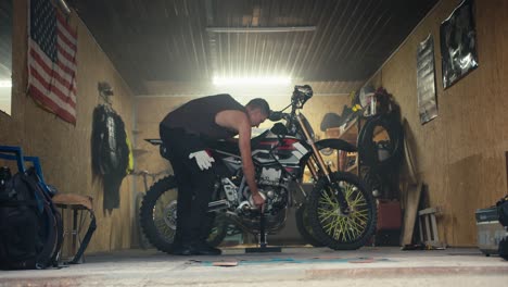 a male mechanic with a short haircut and a gray t-shirt puts his motorcycle on a jack in his garage workshop against of the us flag. start of the working day and start of motorcycle maintenance in the workshop