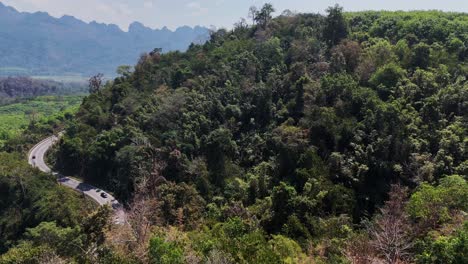 A-bird's-eye-view-of-a-beautiful-road-winding-through-a-forest-in-Songklaburi,-Thailand,-offering-a-scenic-drive-amidst-lush-greenery