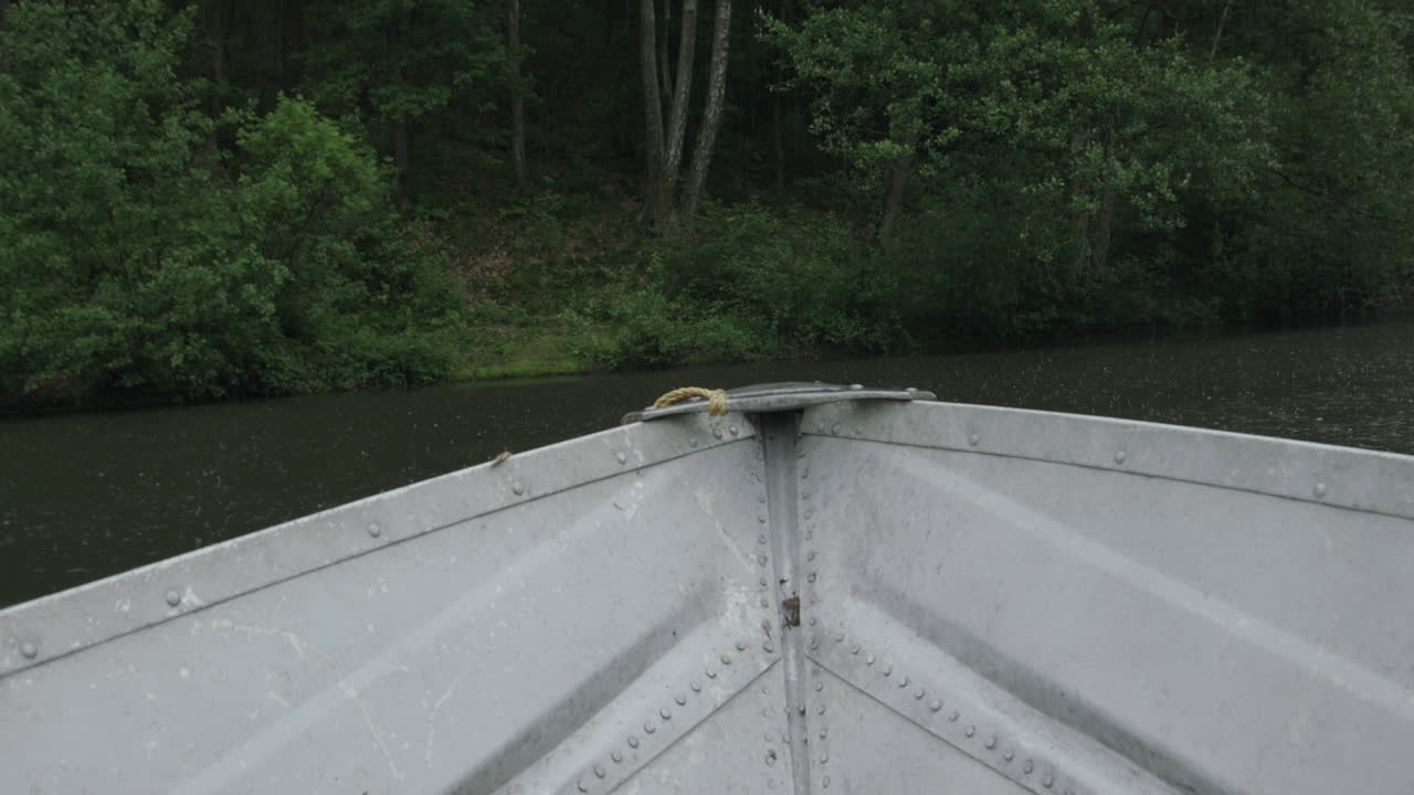 A first-person view from a metal boat navigating a calm river surrounded by dense green forest in heavy rain