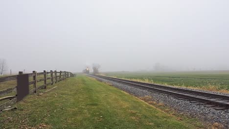 A serene countryside setting reveals a railway track stretching into the fog. A Steam Train Approaches. Lush green fields border the tracks, creating a peaceful yet mysterious atmosphere