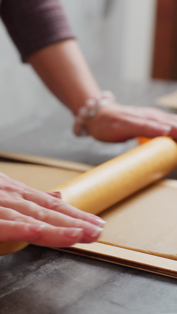 나무 롤링 핀으로 갈색 반죽을 굴리는 여자의 손의 클로즈업, 왼쪽 손목은 분홍색 구슬 팔로 장식되어 있으며, 배경에는 불, 베이킹 도구가있는 약간의 흐릿함이 있습니다.