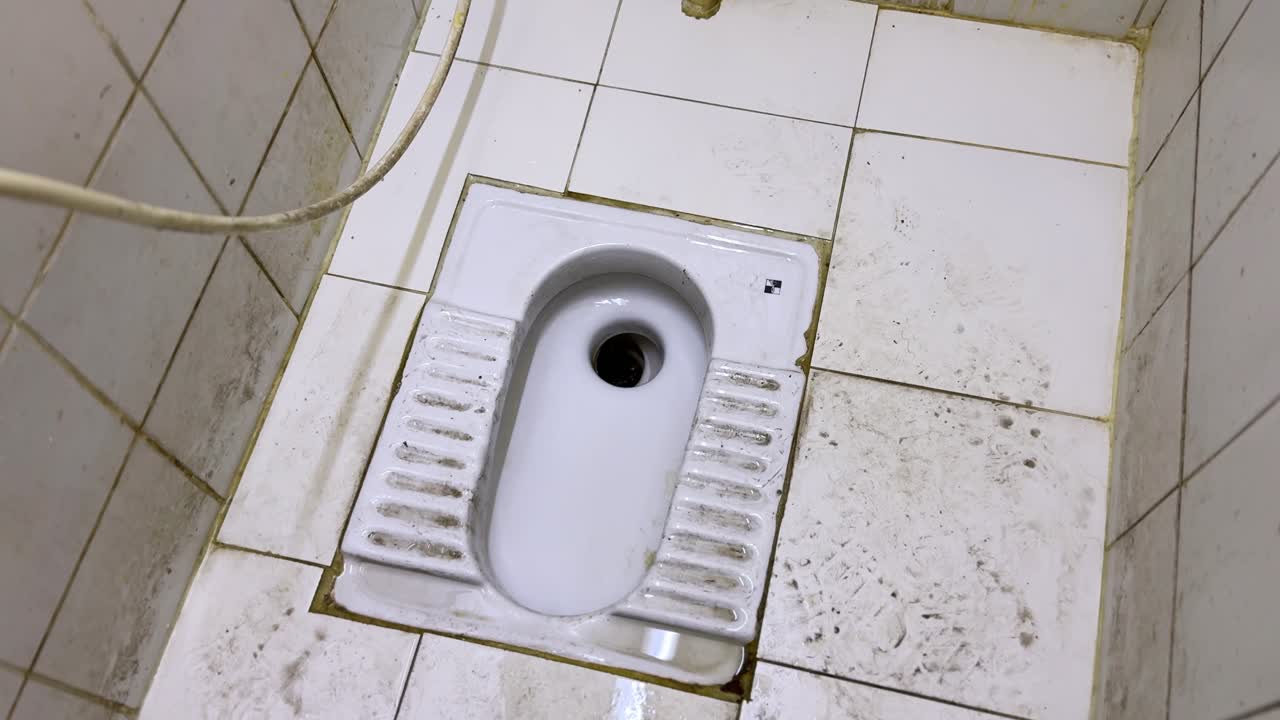 Top view of a ceramic squat toilet with flowing water on a stained tiled restroom floor, showing worn sanitation fixtures and poor hygiene conditions in an indoor washroom