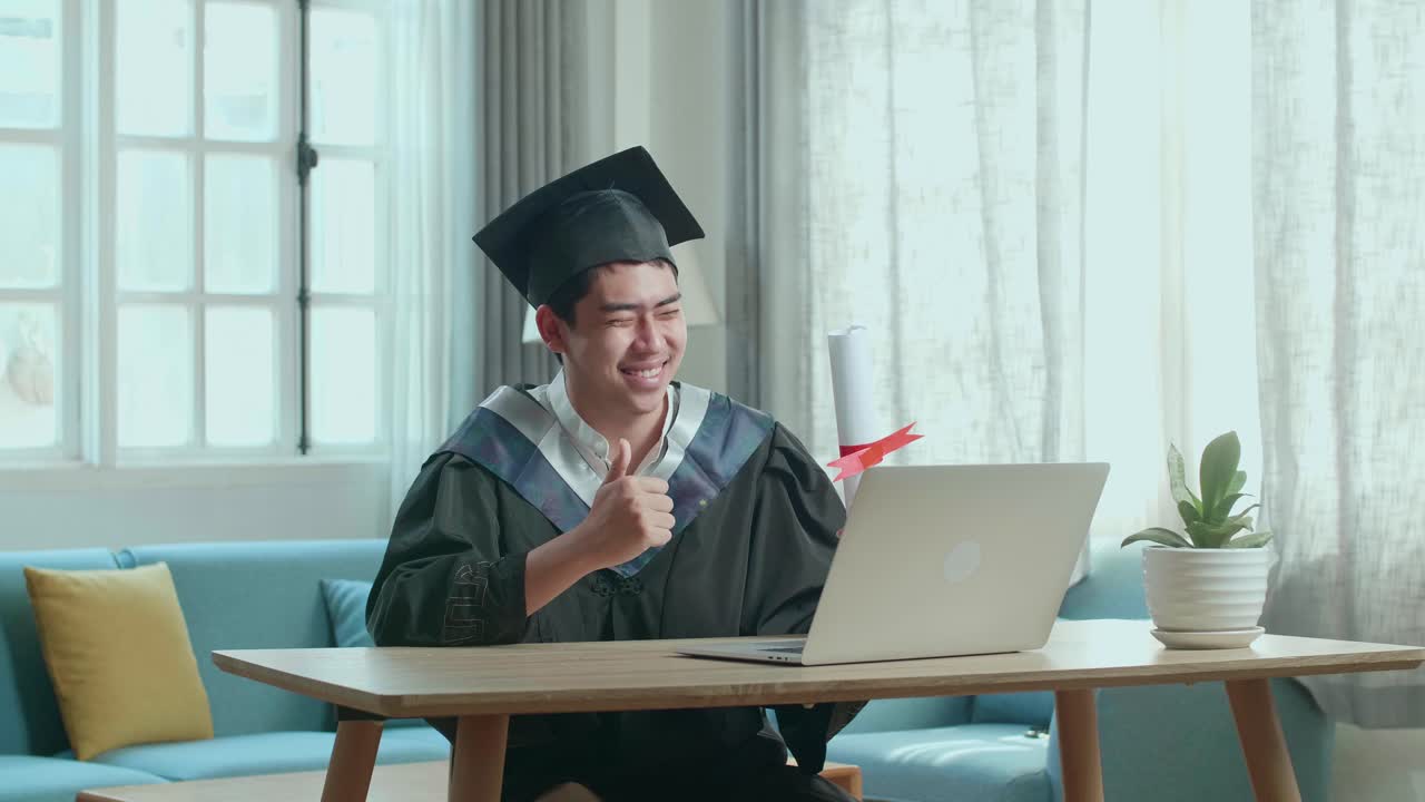 Excited Asian Man Thumb Up And Showing Off A University Certificate To The Family During An Online Video Call. Male Graduate Wearing A Graduation Gown And Cap Sitting On The Living Room