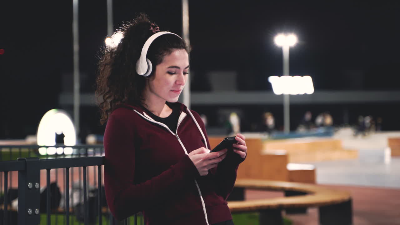 deportista sonriente escuchando música con auriculares bluetooth, enviando mensajes de texto a su teléfono móvil y mirando a su alrededor mientras toma un descanso durante su sesión de entrenamiento por la noche en el parque