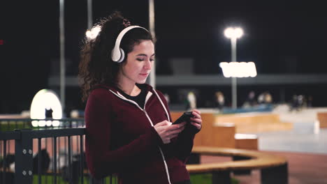 smiling sportswoman listening music with bluetooth headphones, texting on her mobile phone and looking around while taking a break during her training session at night in the park