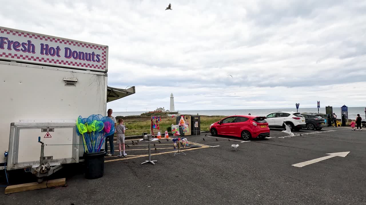 Seagulls flock around a coastal parking lot with a snack van, toy stall, and visible lighthouse under overcast daylight. Camera remains static throughout