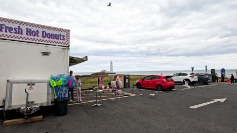 seagulls flock around a coastal parking lot with a snack van, toy stall, and visible lighthouse under overcast daylight. camera remains static throughout