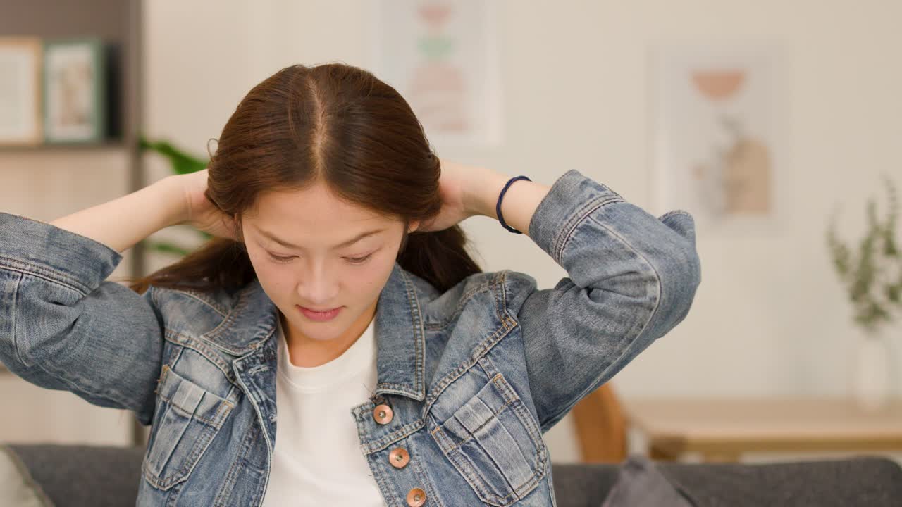 Asian woman casually arranging long hair on living room sofa, soft daylight, static camera