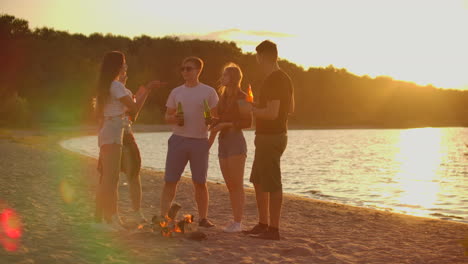 the five students spend time at sunset on the sand beach in shorts and t-shirts around bonfire with beer. they are talking to each other and enjoying the warm summer evening near the river.