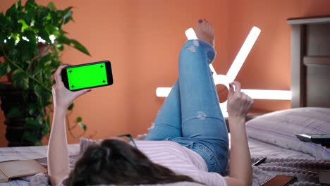 girl holding smartphone with horizontal green screen mock-up, online tracking. female lying on bed and watching video on mobile phone, browsing internet, reading social network news and testing apps