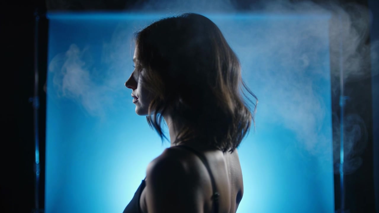 Donna che ruota, con un top nero con spalline sottili, su un set illuminato da dietro di blu, con foschia che si sposta a destra.