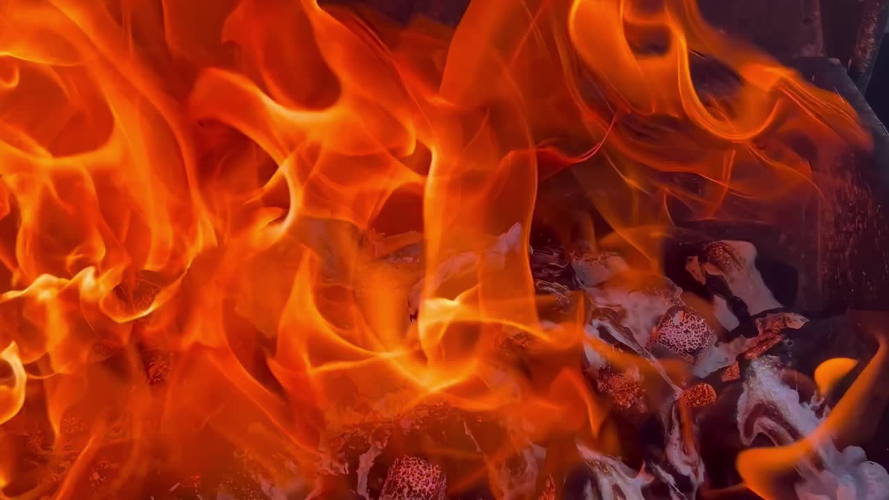 Bright orange flames flicker intensely over glowing coals, creating a mesmerizing display of heat and light.