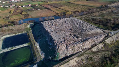 Large-garbage-landfill-on-river-bank,-waste-mountain-and-polluted-water-tanks,-aerial-view