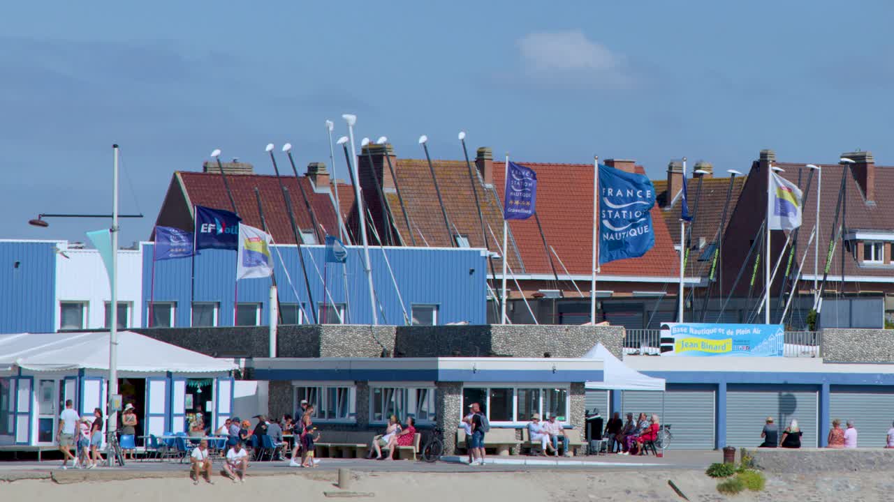 Flags wave in seaside breeze above tourist-filled promenade, sunny day, static wide shot