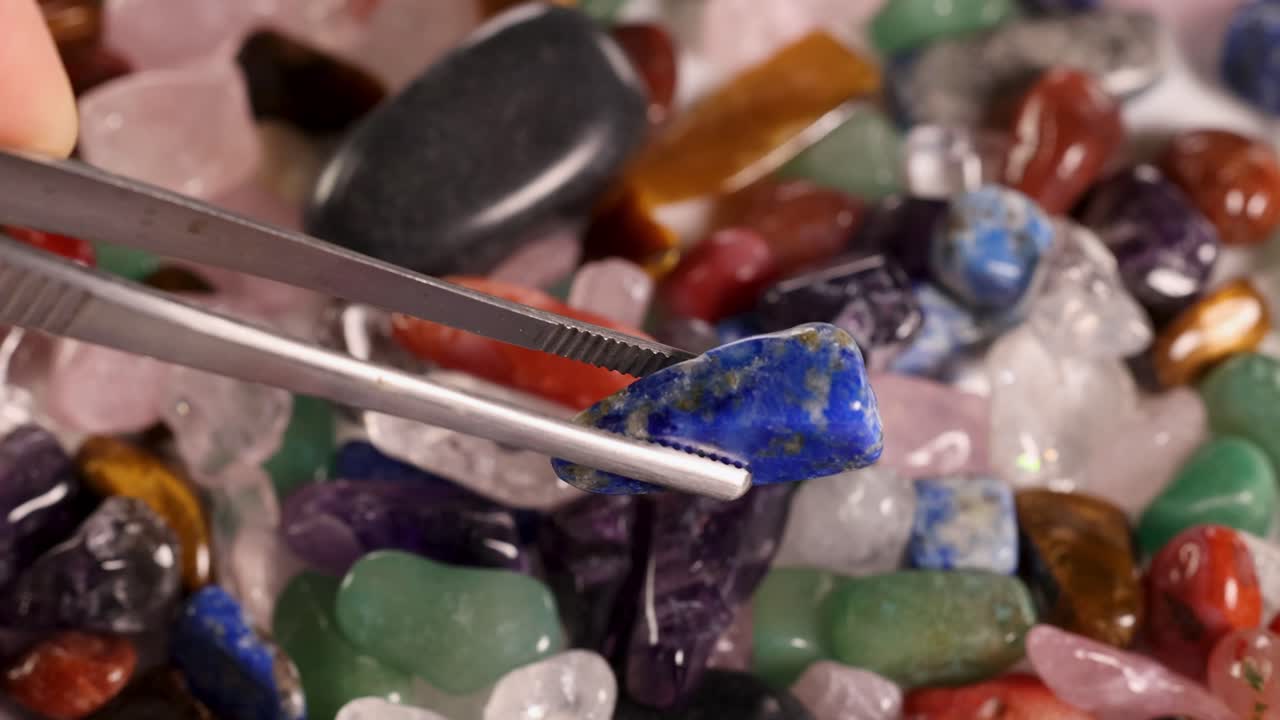 Tweezers carefully pick a lapis lazuli stone from a colorful array of chakra stones under bright lighting