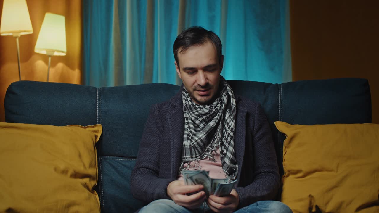 el hombre está sentado en el sofá, sonriendo, contando un paquete de dinero y planeando sus compras. salario del empleado. pago en efectivo. imágenes de 4k