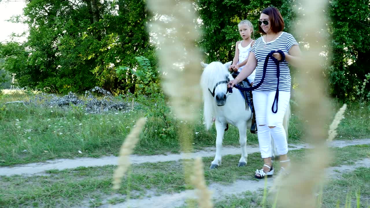 Mom and daughter are walking around the field, daughter is riding a pony, mother is holding a pony for a bridle. Cheerful, happy family vacation. Outdoors, in summer, near the forest