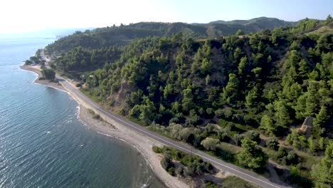 Vista-Aérea-De-Una-Carretera-Junto-A-Una-Playa-Y-Bajo-Una-Colina-Montañosa-En-Grecia