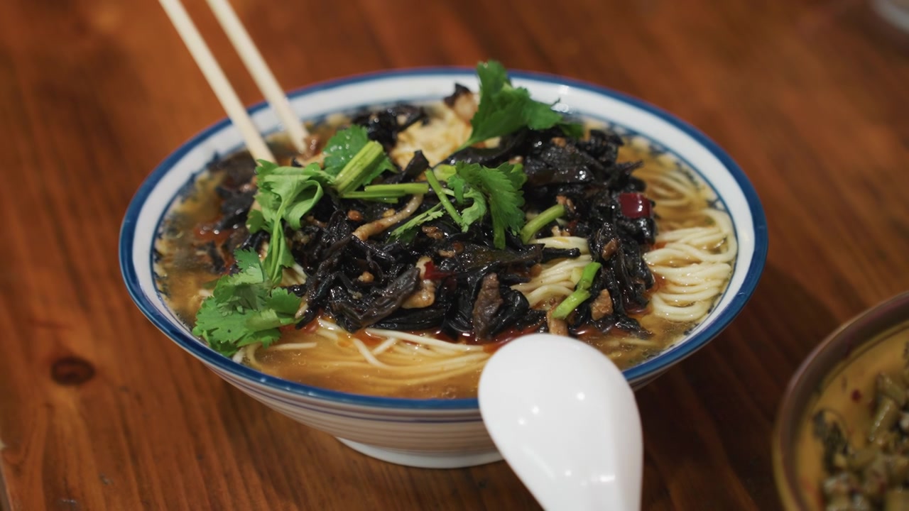 Ricca ciotola di noodles ai funghi su tavolo di legno, brodo fumante guarnito con funghi orecchio di Giuda e coriandolo fresco, bacchette che sollevano fili di noodles, cucchiaio di porcellana appoggiato sul bordo, illuminazione calda color ambra