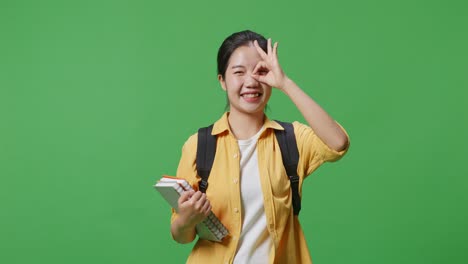 asian woman student with a backpack and some books smiling and showing okay hand sign over eye while standing in the green screen background studio