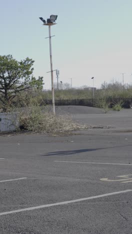 vertical video of abandoned car park with weeds growing from decaying tarmac and disused flood lights with metal fence and trees in background. post apocalyptic setting.