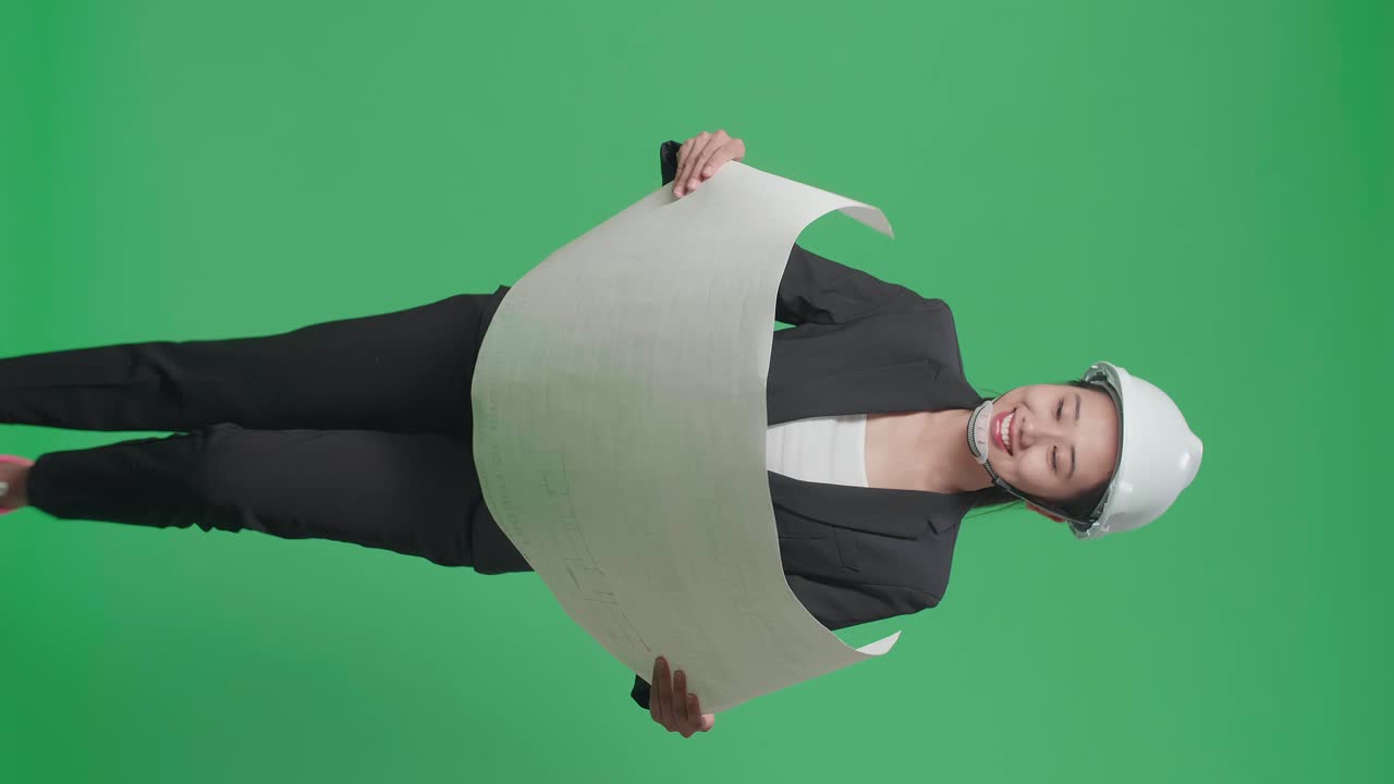 Smiling Asian Female Engineer With Safety Helmet Looking At Blueprint While Walking In The Green Screen Studio