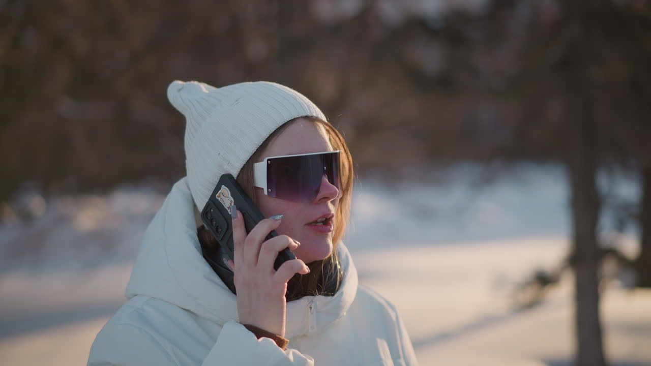Young woman holds phone to ear singing and dancing in rhythm under sunset light wearing beanie and puffer coat near statue in park with snow and trees blurred in background conveying joyful vibe