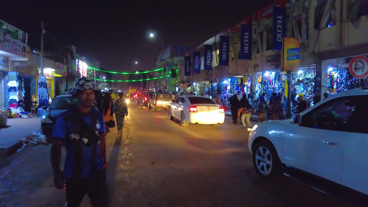 Handheld shot of a busy street in the middle of serekunda market, gambia overlooking the dense slow traffic with shops, pedestrians and colourful street lights during a beautiful warm night