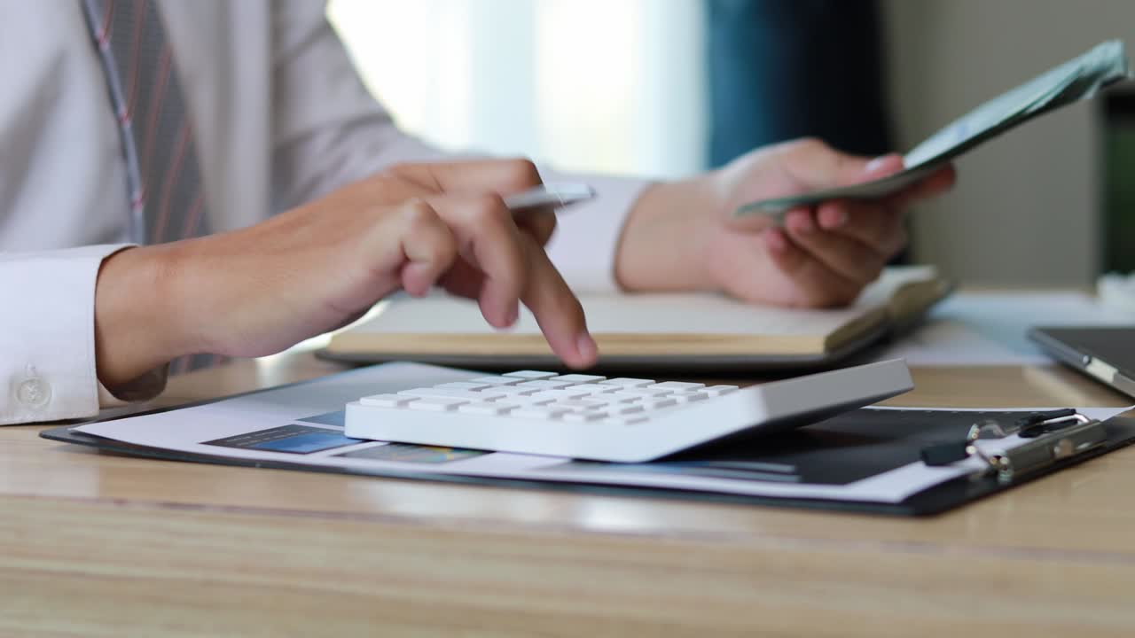 un hombre de negocios usando su mano derecha para presionar una calculadora, su mano izquierda sosteniendo un dólar, está calculando el dinero. metraje 4k.