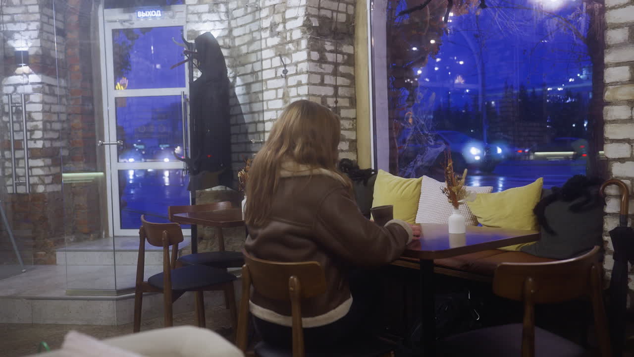 Young girl in brown shearling jacket holding coffee cup, walking toward chair, and sitting down at cozy cafe. The soft lighting and ambiance create a warm, inviting atmosphere