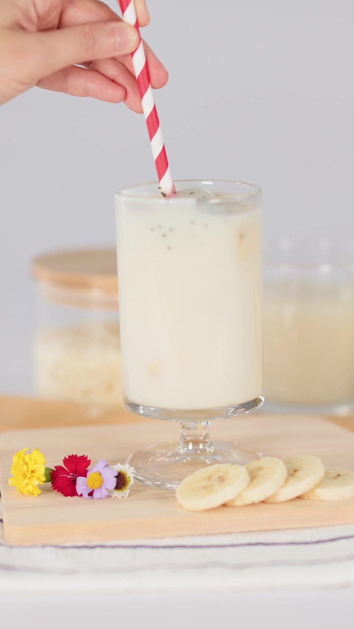 Una mano revuelve un batido cremoso de plátano en un vaso con una pajita a rayas sobre una mesa luminosa.