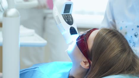 a doctor in white latex gloves turns on a laser tooth whitening device. the girl at the dentist in red protective uv glasses