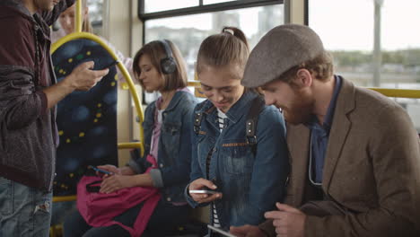 joven con barba y boina usando un teléfono inteligente mientras habla con una joven sosteniendo un teléfono inteligente, ambos viajando en autobús en la ciudad