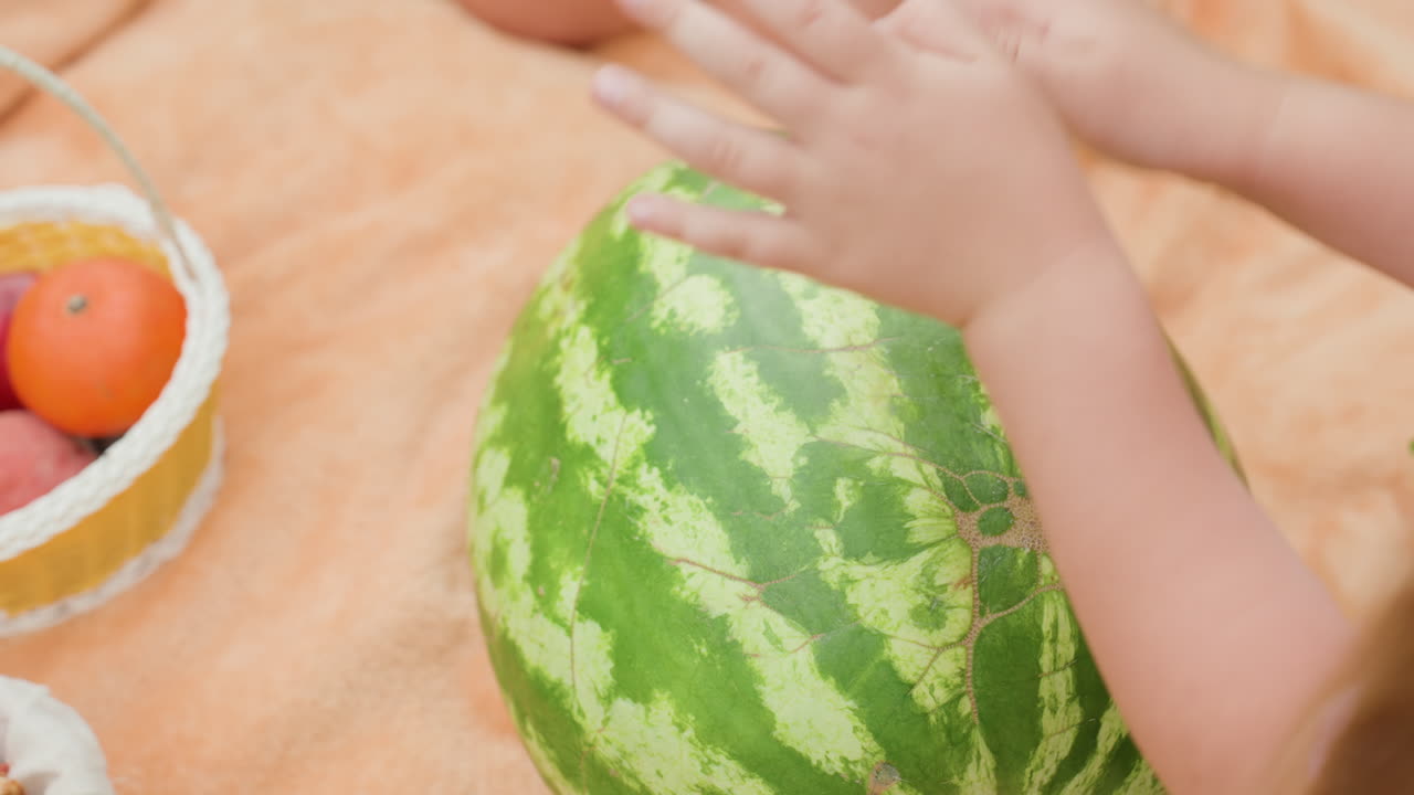 과일 바구니 근처에서 흰색 옷을 입은 소녀가 수박 껍질을 두드리고 있어요. 금발 머리가 보이고, 햇빛이 비치는 분위기 속에서 수박이 잘 익었는지 확인하려고 귀를 기울이고 있어요. 손이 클로즈업된 모습에서 여름을 기대하는 장난기 넘치는 시장 피크닉 분위기가 느껴져요.