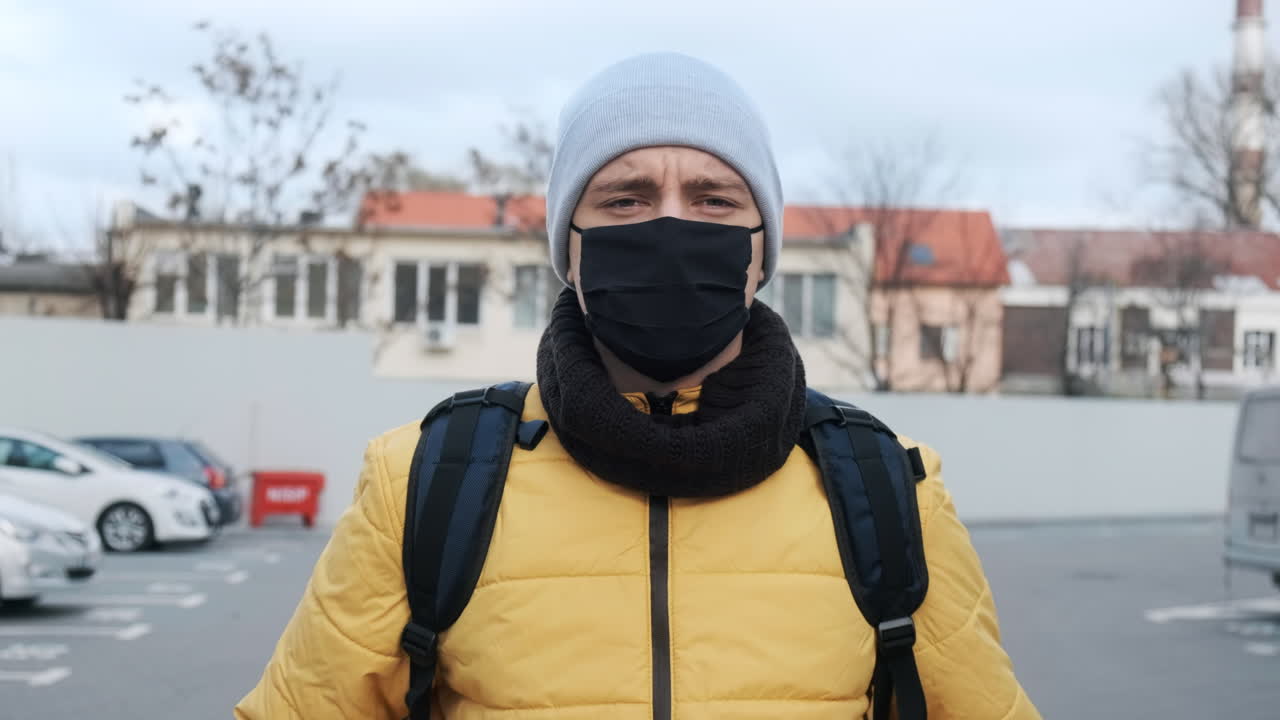 Food courier with backpack and black medical mask on parking, watching into camera. Food delivery service